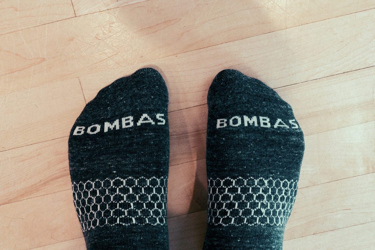 A pair of feet in charcoal Bombas socks with BOMBAS knitted across the toes and the brand’s honeycomb arch pattern, standing on a light wood floor