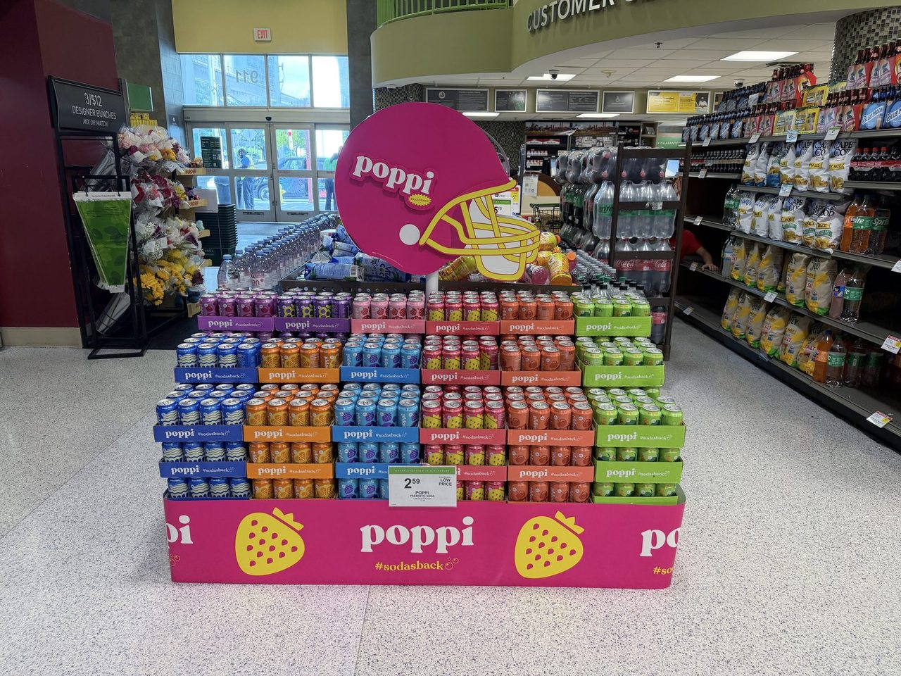 A supermarket end-cap display stacked with cases of Poppi prebiotic soda in assorted flavors under a pink Poppi football-helmet sign