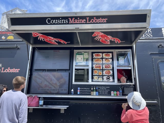 A black Cousins Maine Lobster food truck with red lobster graphics and a lit menu window, with customers lined up at the counter on a sunny day in the San Francisco Bay Area