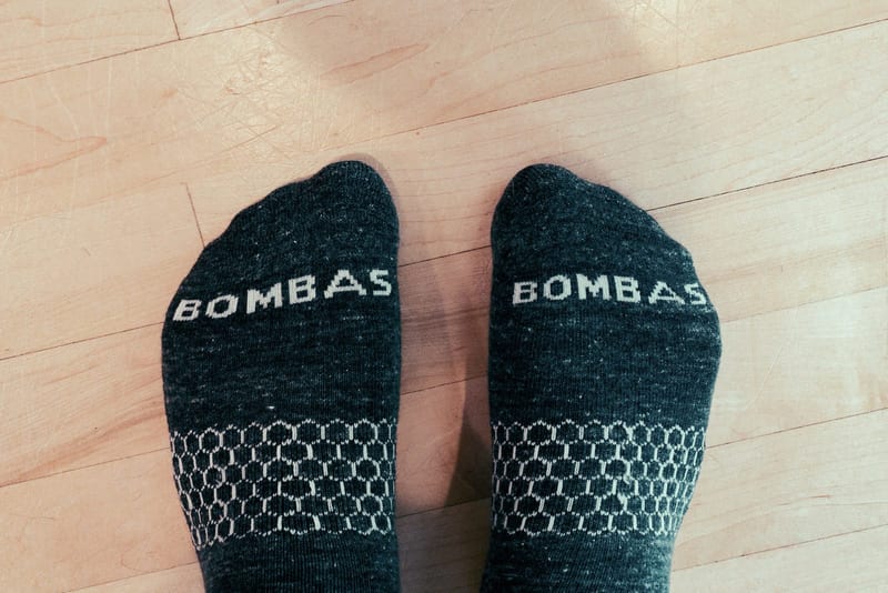 A pair of feet in charcoal Bombas socks with BOMBAS knitted across the toes and the brand’s honeycomb arch pattern, standing on a light wood floor