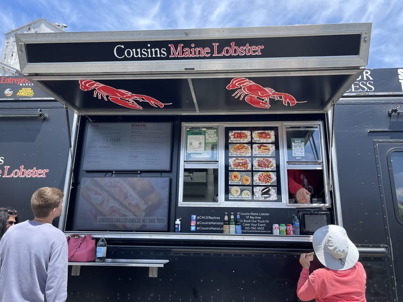 A black Cousins Maine Lobster food truck with red lobster graphics and a lit menu window, with customers lined up at the counter on a sunny day in the San Francisco Bay Area