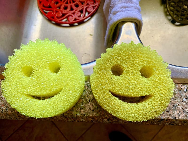 Two yellow smiley-face Scrub Daddy sponges propped on the edge of a stainless-steel kitchen sink