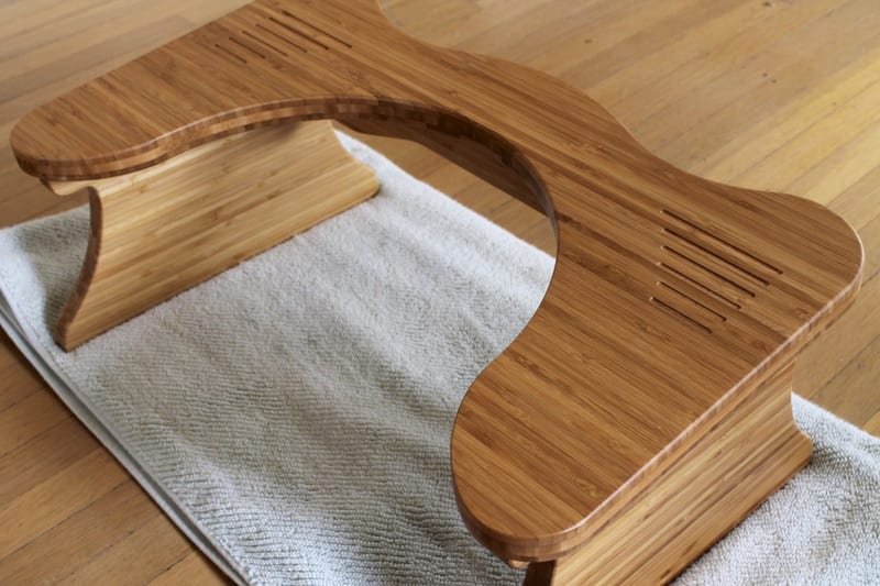 A curved bamboo Squatty Potty toilet stool resting on a white bath mat on a wooden floor